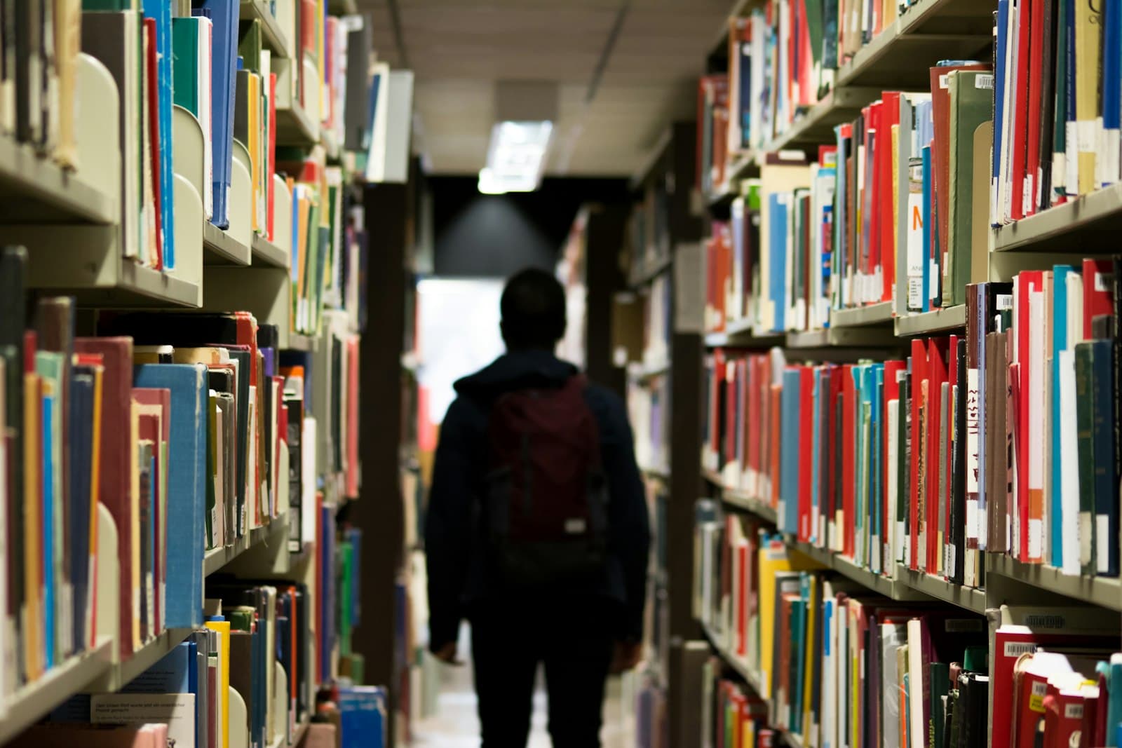A student walking down a library aisle between tall bookshelves